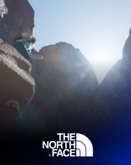 A woman jumps across 2 big rocks on a hike, with The North Face white logo on top of the image. 